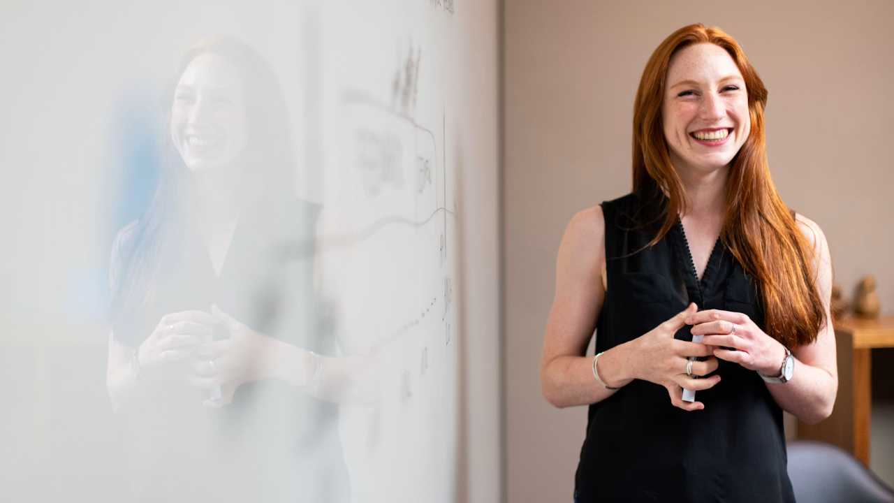 Professional-Woman-Standing-By-Whiteboard A woman with red hair wearing a black shirt stands with a marker in her hand next to a whiteboard.