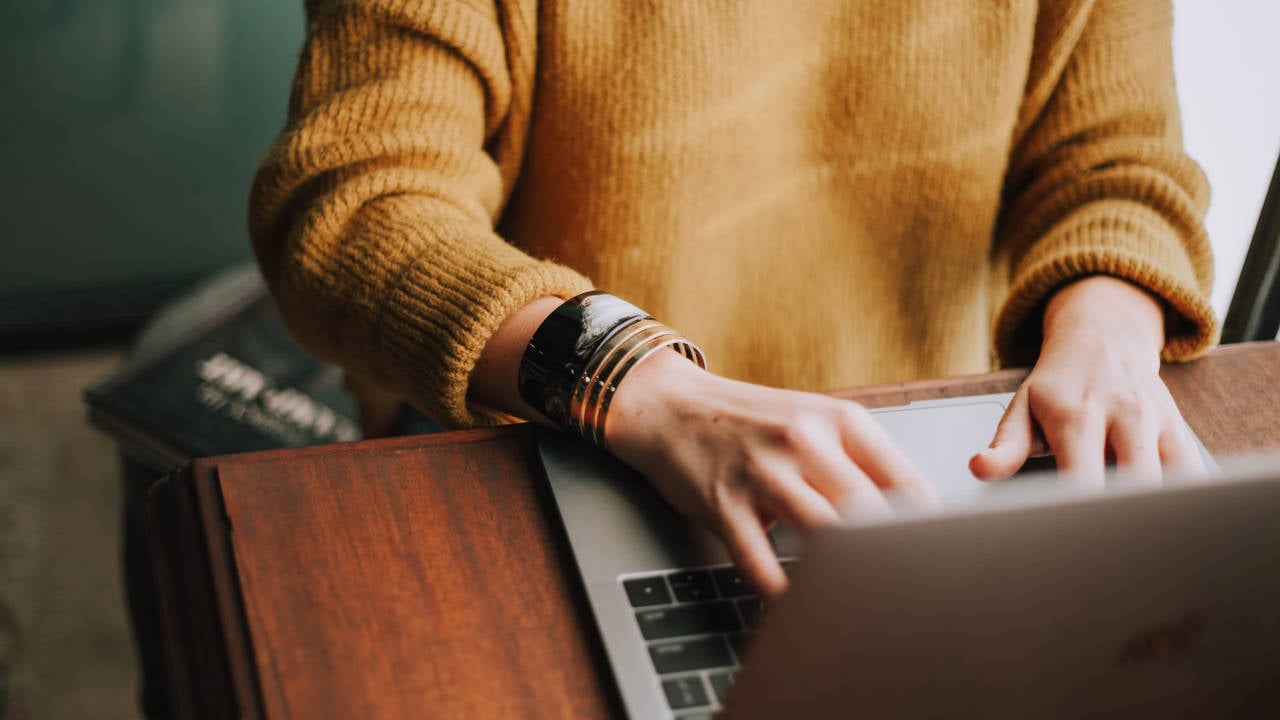A small business owner wearing a yellow sweater working on a MacBook at a wooden desk in a green room.